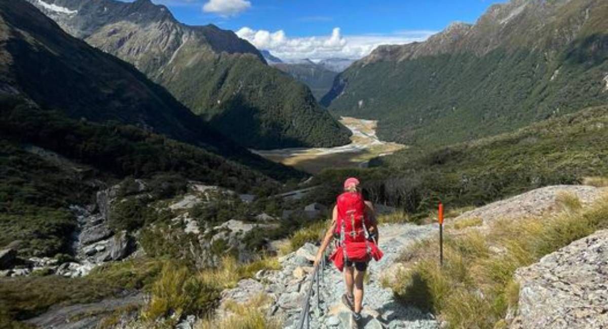 A hiker in red attire walking on the Routeburn Track, surrounded by majestic mountains and a lush green valley.